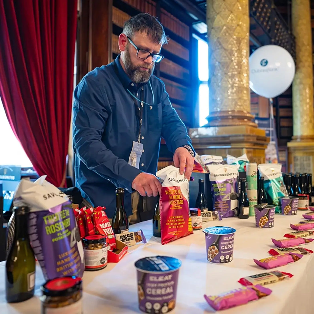 A man in a blue shirt and glasses stands behind a table covered with packaged food products, including bags and tubs of cereal and snack items, in an indoor event space with red curtains and decorative columns.