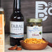 FABAL Lager bottle, MANI Olives jar, and bowl of snacks on a wooden surface with a blurred background.