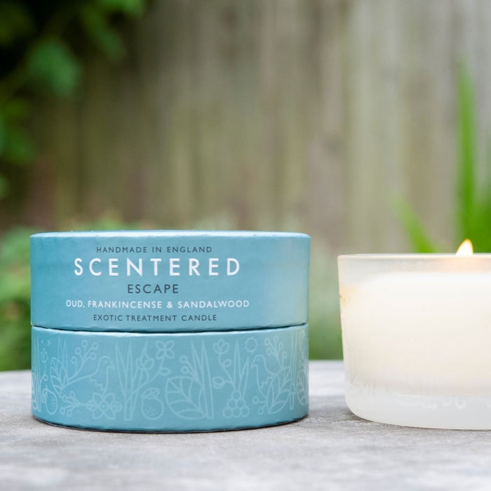 Scentered Escape Relaxation Travel Candle 85g alongside the Escape Travel Candle coloured round box with plants in background