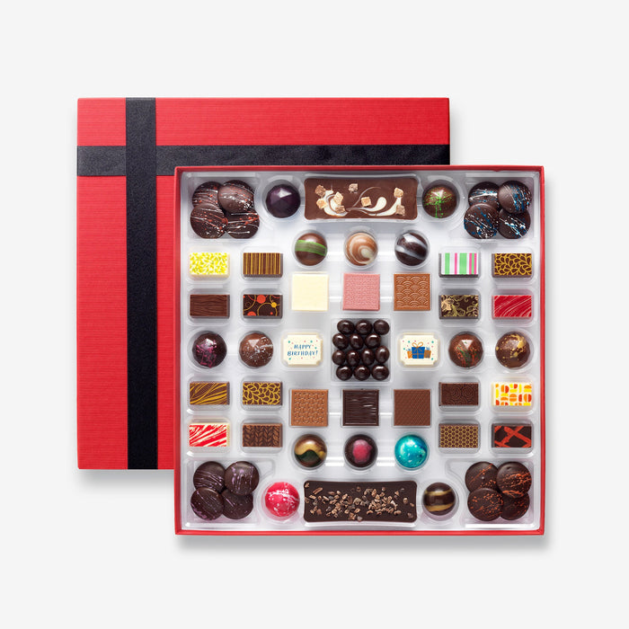 An open chocolate selection box containing 55 chocolates made by Harry Specters. The chocolates seen within this gift box are a colourful mix of white, milk, ruby, caramel, and dark chocolate with coffee beans, mini chocolate bars, and hot chocolate buttons with two Happy Birthday themed chocolates.